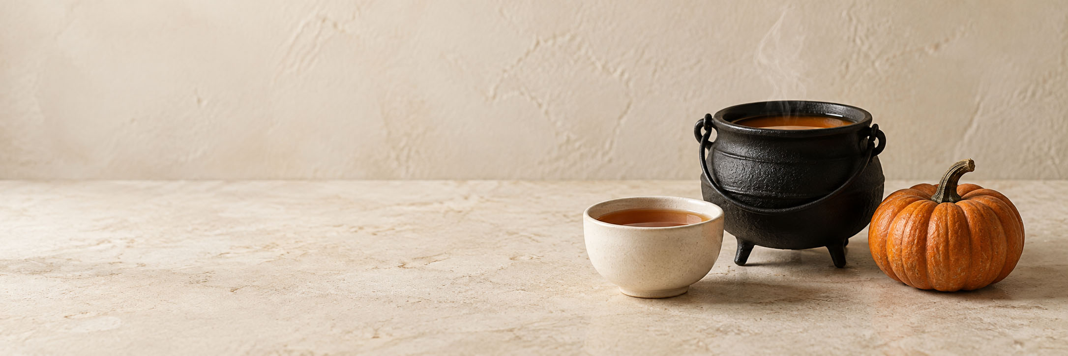 A small black cauldron and ceramic cup of amber tea beside a pumpkin on a quiet cream tabletop